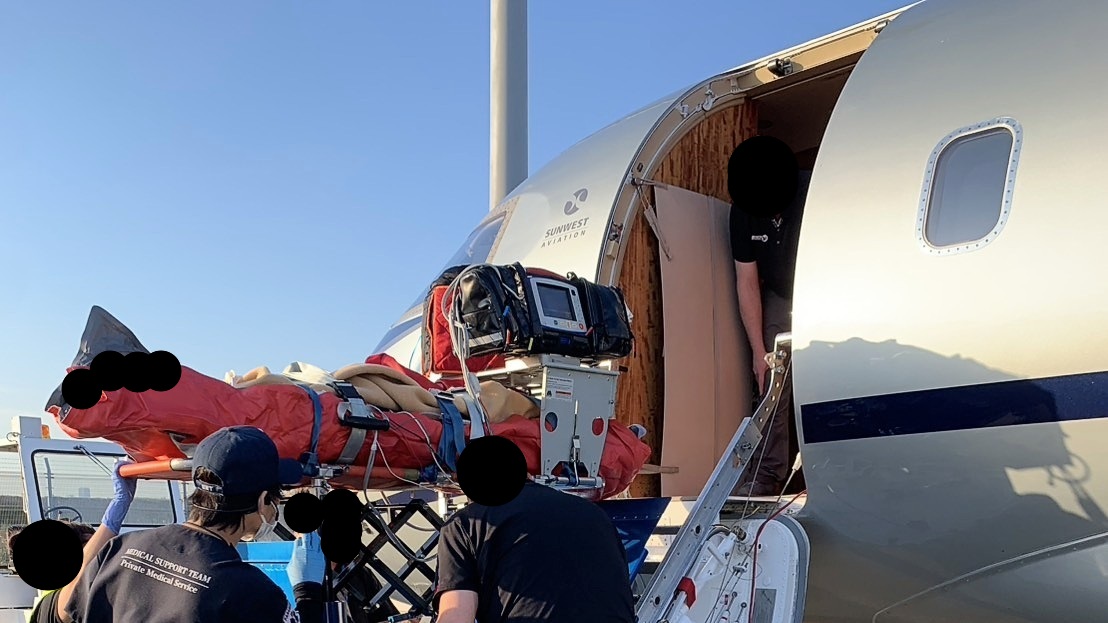 晴天の空港滑走路で、搬送車とエアアンビランス機、機体近くのストレッチャーが写る医療フライト搬送準備の様子